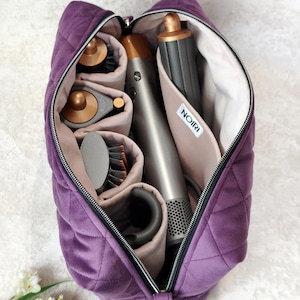 Pode incluir: Uma bolsa de armazenamento de veludo roxo acolchoado com fecho de correr, contendo uma ferramenta de modelagem de cabelo cinzenta e vários acessórios de cor cobre e cinzento organizados em bolsos internos. Uma etiqueta branca com o texto NOIRI é visível.