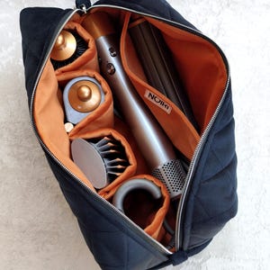 May include: A black quilted travel case with a zipper closure and a vibrant orange interior lining. The case features internal dividers holding a silver hair styling tool with gold accents and several circular attachments, with a white tag labeled NOIRI.