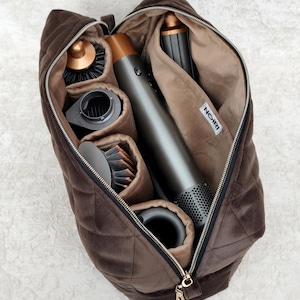 May include: A dark brown quilted velvet storage bag with a zipper closure, containing a gray and copper hair styling tool with multiple attachments. The interior is lined with light brown fabric and features a white tag labeled NOIRI.