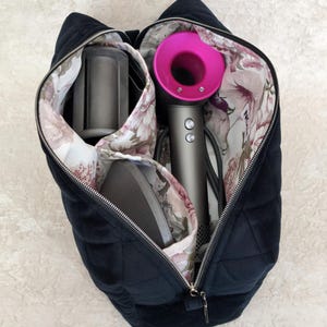 Pode incluir: Uma bolsa de armazenamento preta acolchoada com zíper e forro interno floral contendo um secador de cabelo cinza com bocal circular rosa brilhante e dois acessórios de modelagem cinzas. A bolsa possui um formato estruturado.