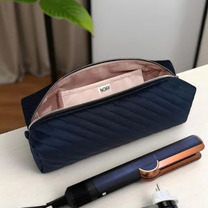 Könnte beinhalten: Eine marineblaue, gesteppte Kosmetiktasche mit einem passenden kleinen Beutel im Inneren, der mit "NOIRI" beschriftet ist. Ein blau-kupferfarbenes Glätteisen befindet sich neben der Tasche. Die Tasche ist geöffnet und zeigt ein rosa Innenfutter.