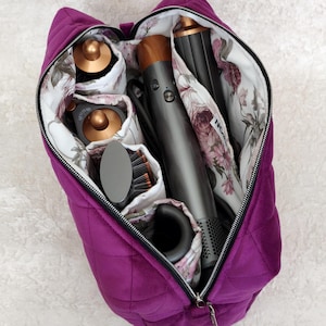 May include: A quilted deep purple storage bag with a zipper closure, featuring an interior lined with a white fabric printed with pink and green floral patterns. The bag contains a gray hair styling tool with copper accents and attachments.