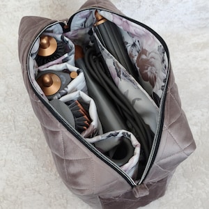 May include: A quilted taupe velvet storage bag with a black zipper closure. The interior is lined with a white fabric featuring a floral pattern in shades of pink and gray. It contains various hair styling tools with copper and gray accents.
