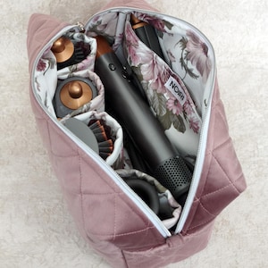 May include: A dusty rose quilted velvet storage bag with a white interior lining featuring a floral pattern. The bag contains a dark gray hair styling tool and several attachments. A white label with the black text NOIRI is visible inside.
