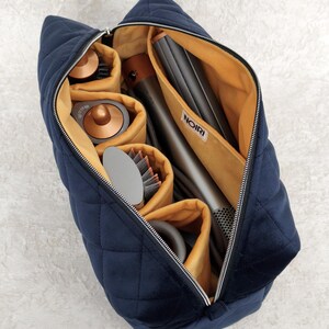 May include: A navy blue quilted velvet travel bag with a zipper closure and a mustard yellow interior lining. The bag features internal compartments containing various hair styling tools in metallic copper and gray tones, with a white label reading NOIRI.