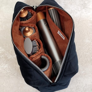 Pode incluir: Um estojo de viagem acolchoado preto com fecho de correr e forro interior macio na cor ferrugem. O estojo contém uma ferramenta de modelagem de cabelo em cinzento metálico e cobre, juntamente com vários acessórios e escovas a condizer.