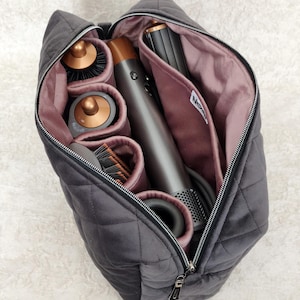 May include: A charcoal gray quilted storage bag with a zippered opening and a mauve interior lining. The bag contains a hair styling tool with copper accents, multiple cylindrical attachments, and a small white label with black text inside.