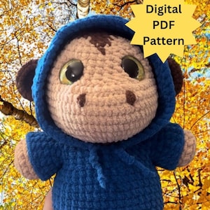 May include: A crocheted plush toy wearing a blue hooded sweater. The toy has brown ears, a brown nose, and large brown eyes. The toy is being held in a hand against a background of autumn foliage. The text "Digital PDF Pattern" is in a yellow starburst shape above the toy. The text "@mamas.lil.minis" is below the toy.