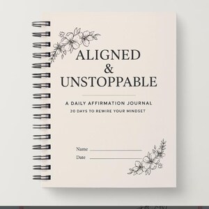 May include: A spiral-bound journal with a cream-colored cover. The cover features the words "ALIGNED & UNSTOPPABLE" in black, along with floral illustrations. The text "A DAILY AFFIRMATION JOURNAL 20 DAYS TO REWIRE YOUR MINDSET" is also visible.