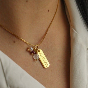 Puede incluir: Un collar de cadena dorada con un dije que dice "BEAUTIFUL" y tiene pequeñas cuentas rosadas, blancas y transparentes adjuntas.