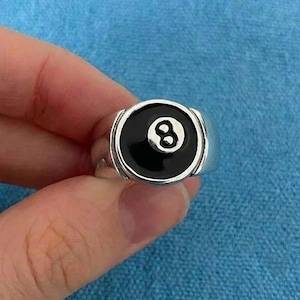 May include: A silver ring with a black enamel face displaying the number 8 in white, styled after a billiard eight ball. The ring has a rounded rectangular shape and a polished finish, suitable for casual wear.