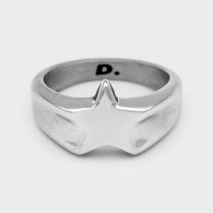 Könnte beinhalten: Ein silberfarbener Metallring mit einem markanten sternförmigen Design auf der Vorderseite. Die Innenseite des Rings ist deutlich mit dem Buchstaben D gefolgt von einem Punkt graviert, der auf der glatten, polierten Metalloberfläche sichtbar ist.
