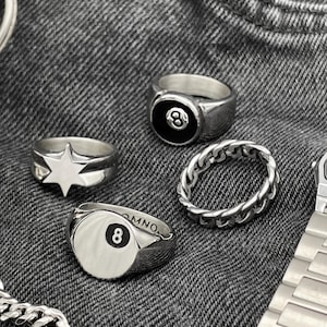 May include: Four silver rings with different designs. One ring features a black and white eight ball, another ring has a star design, and another ring has a silver eight ball design. The fourth ring is a silver chain link design.