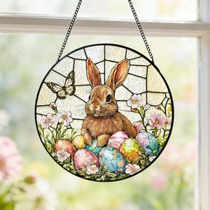 Peut inclure: Vitrail rond représentant un lapin brun entouré d'œufs de Pâques colorés et de fleurs. Un papillon est également présent. La fenêtre est suspendue par une chaîne noire. L'œuvre est placée devant une fenêtre.