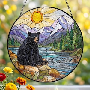 Könnte beinhalten: Ein runder Buntglas-Sonnenfänger mit einem schwarzen Bären, der auf Felsen an einem Fluss sitzt. Die Szene zeigt eine leuchtend gelbe Sonne, violette Berge und grüne Tannen. Das Stück ist in schwarzes Metall gefasst und hängt an einer Kette.