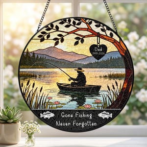 May include: A circular hanging suncatcher featuring a silhouette of a person fishing in a boat on a lake with mountains in the background. A heart-shaped ornament hangs from a tree branch with the text Dad 1949-2026. Text reads Gone Fishing Never Forgotten.