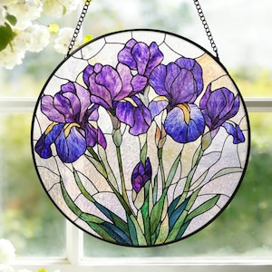 Op de afbeelding: Een rond gebrandschilderd glazen raam met paarse en blauwe irissen. De bloemen hebben gedetailleerde bloemblaadjes en groene stelen en bladeren. Het raam is zwart omlijst en hangt aan een ketting.