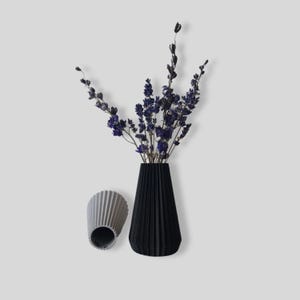 May include: A black and gray pleated vase with dried purple flowers. The vase is sitting on a white surface.
