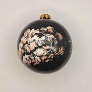 Può includere: Una palla di Natale nera con un cappuccio dorato e un disegno dipinto a forma di nuvola nei colori bianco e pesca. La palla è rotonda e ha una finitura lucida.