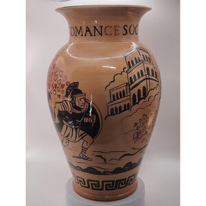 May include: A tan ceramic vase with black and red illustrations. The vase features a Roman soldier, hearts, and a building. The words "ROMANCE SOC" are printed on the top of the vase. The vase has a Greek key design around the base.