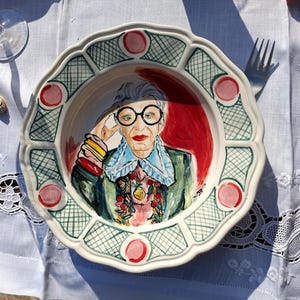 Può includere: Un piatto decorativo in ceramica con il ritratto di una donna con occhiali rotondi, rossetto rosso e un abito colorato. Il piatto ha un bordo smerlato con accenti verdi e rosa.
