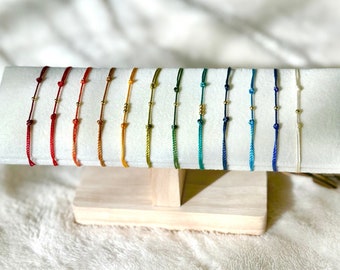 Könnte beinhalten: Eine Präsentation farbenfroher Schnurarmbänder, jedes mit einer kleinen goldenen Perle. Die Armbänder sind in Regenbogenfarben angeordnet, von Rot bis Blau, und werden auf einem weißen, rechteckigen Ständer mit Holzsockel präsentiert. Der Hintergrund ist eine weiche, weiße, strukturierte Oberfläche.