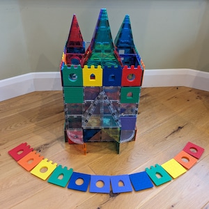 Puede incluir: Un castillo de baldosas magnéticas de colores con agujas transparentes rojas, verdes y azules. El castillo tiene bloques cuadrados y rectangulares en varios colores. Un arco iris de bloques de espuma rodea el castillo.