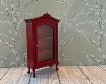 May include: A miniature red display cabinet with a glass door and shelves. The cabinet has a curved top and sits on four curved legs. The background features a light green wall with a white floral pattern, and a wooden floor.