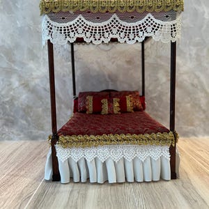 Pode incluir: Cama com dossel em miniatura com estrutura de madeira escura. A cama apresenta um edredom e almofadas estampadas em vermelho, acentuadas com acabamentos dourados. Renda branca e acabamentos dourados adornam o dossel e a saia da cama. Uma peça decorativa.