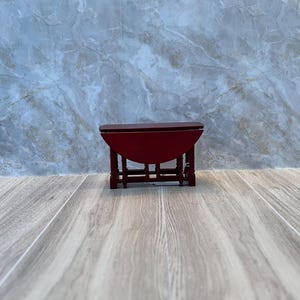 May include: A small, red wooden drop-leaf table. The table has a rounded top and a curved apron. The legs are straight and connected by a stretcher. The table is approximately 12 inches long when closed.