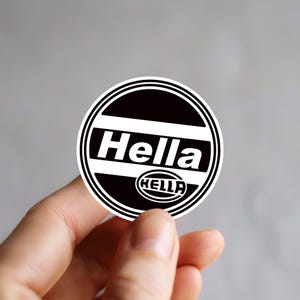 Peut inclure: Un autocollant circulaire présentant un design noir et blanc avec le mot Hella dans une police sans empattement en gras au centre. Sous le texte principal, un petit emblème ovale contient également le mot Hella dans une police stylisée.