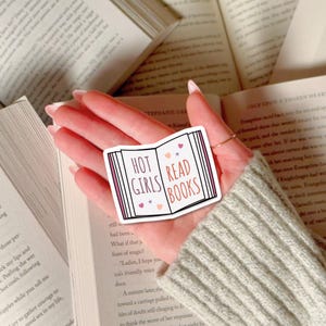 May include: A sticker with a pink and white book design. The book has the words "Hot Girls Read Books" on the cover. The sticker is held in a hand over a stack of books.