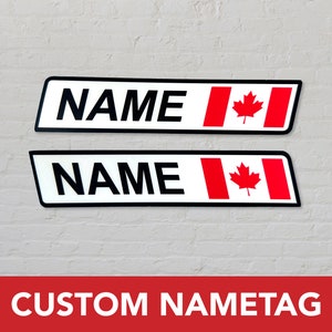 Peut inclure: Deux étiquettes de nom rectangulaires blanches avec du texte noir et un motif de drapeau canadien. Le texte sur les étiquettes de nom dit "NAME". Les étiquettes de nom sont sur un mur de briques blanches. Le texte en bas de l'image dit "CUSTOM NAMETAG".