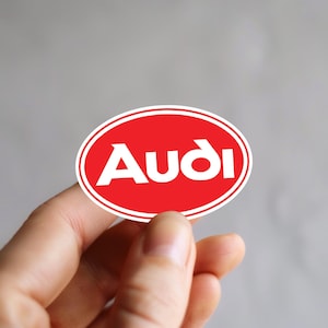 Könnte beinhalten: Ein roter ovaler Aufkleber mit weißem Rand und dem weißen Wort "Audi" in fetter Schrift. Der Aufkleber wird zwischen Fingern vor grauem Hintergrund gehalten.