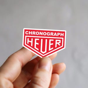 May include: A red and white sticker with the word "CHRONOGRAPH" at the top and "HEUER" in bold letters in the center. The sticker has a white border and is held by a hand.