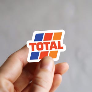 May include: A sticker featuring the word "TOTAL" in red, with a stylized logo of blue, orange, and red squares. The sticker is held between fingers against a gray background.