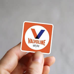 May include: A square sticker with the Valvoline logo. The logo has a white circle with a red and blue V-shaped design, the word "VALVOLINE" in red, and two checkered flags. The sticker is set against an orange background.