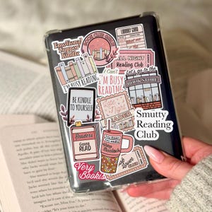 Peut inclure: Liseuse noire dans un étui transparent, ornée d'autocollants. Les autocollants affichent des phrases comme "Smutty Reading Club" et "Readers Gonna Read". La liseuse est tenue au-dessus d'un livre ouvert.