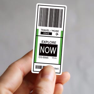 Peut inclure: Un autocollant blanc en forme de billet de voyage, tenu par une main. Il présente des lignes de code-barres noires, le texte "TRAVEL / MORE 0365" avec une icône d'avion, et les mots "EXPLORE NOW" en gras. La date "05JUL" est également visible.