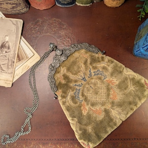 Peut inclure: Un sac à main ancien en velours vert olive avec un cadre et une chaîne en métal argenté. Le sac présente un motif floral brodé dans des tons orange et bleu. Des photographies anciennes sont en arrière-plan.