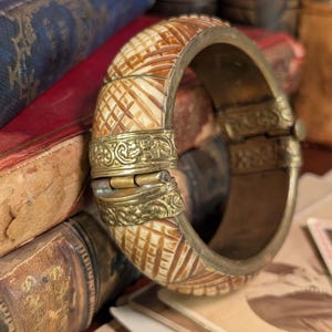 May include: An antique hinged bracelet with a cream and brown patterned design. The bracelet has brass accents with ornate detailing and a secure clasp. The bracelet is displayed on a stack of old books with red and blue covers.