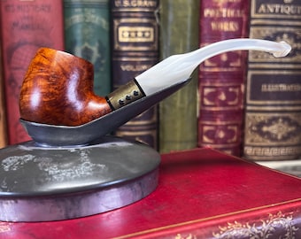 May include: A handcrafted tobacco pipe with a brown bowl and a white stem, resting on a metal stand. The pipe features a detailed design and is set against a backdrop of antique books, suggesting a vintage aesthetic.