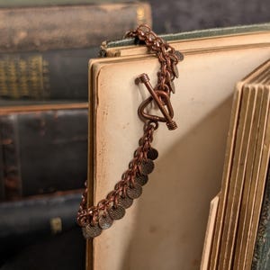 Può includere: Un bracciale a catena color rame caratterizzato da una serie di piccoli dischi metallici circolari e testurizzati fissati alle maglie. Il bracciale è fissato con una chiusura a T composta da una barra a T e un anello triangolare.
