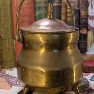 May include: A small brass pot with a lid and a handle. The pot has a rounded body and three legs. It is sitting on a surface with a butterfly pattern.