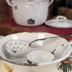 May include: A collection of vintage white enamelware kitchen items. Includes a large oval dish with handles, a small colander, a ladle, and a lidded pot. The enamelware has a distressed, rustic appearance with black trim and visible wear.