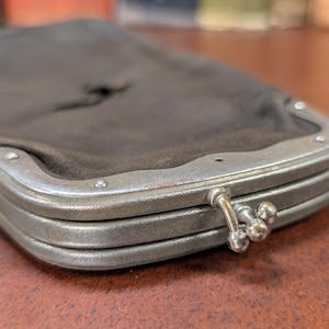 Op de afbeelding: Close-up van een vintage donkerbruine clutch portemonnee met een zilverkleurig metalen frame. De portemonnee heeft een sluiting met een sluiting en is gemaakt van een zacht materiaal. Het metalen frame is aan de portemonnee geklonken.