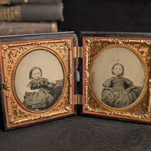 May include: A double-sided daguerreotype case featuring two oval portraits of children in dark dresses. The portraits are framed by ornate, embossed gold-colored metal mats with intricate scrollwork, set within a black hinged case.