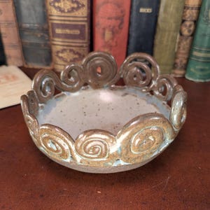 May include: A handcrafted ceramic bowl with a unique, swirling rim design. The bowl features a speckled, light blue interior and a brown, textured exterior. The decorative rim has a spiral pattern. This pottery bowl is ideal for serving or display.