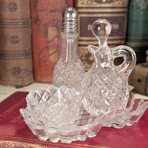 May include: A set of three clear glass bottles with a diamond pattern. The bottles are on a clear glass tray with a scalloped edge. The bottles are for salt, pepper, and oil.