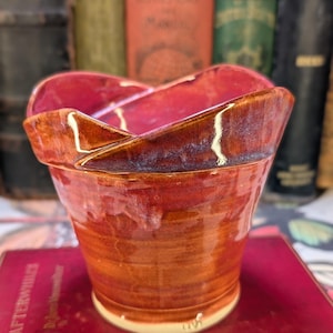 May include: A hand-crafted, red and brown ceramic planter with a unique, folded design. The planter has a glossy finish and a slightly uneven rim.